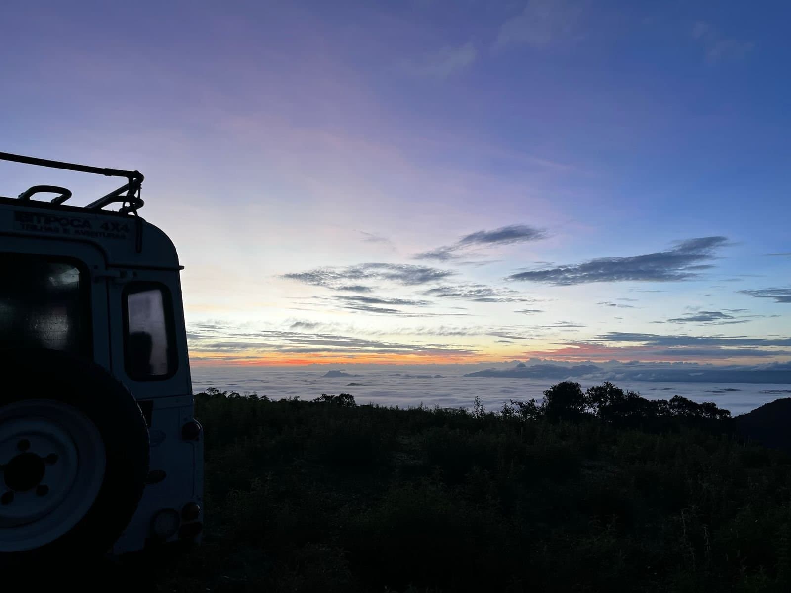 Up before the valley, parked above the clouds. Not a bad start. A 4x4 expedition vehicle silhouetted at dawn above a sea of clouds, with a warm orange and blue horizon stretching across the highland reserve of the Ibiti Project in Minas Gerais