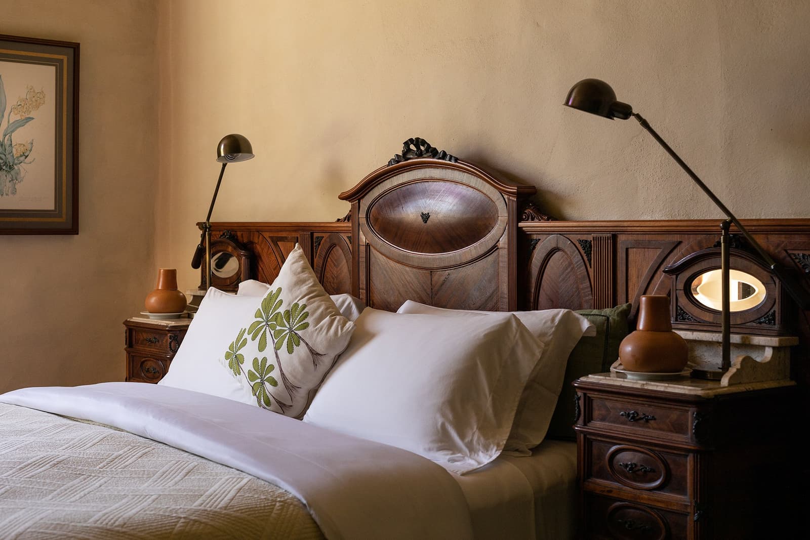 Antique bones, fresh linen, and a pillow that knows its botany. Photo: Henrique Mangeon A double bed with crisp white linen and a botanical embroidered pillow sits against an ornate carved walnut headboard at the Ibiti Project's Engenho Lodge, flanked by matching antique wooden nightstands and warm brass reading lamps