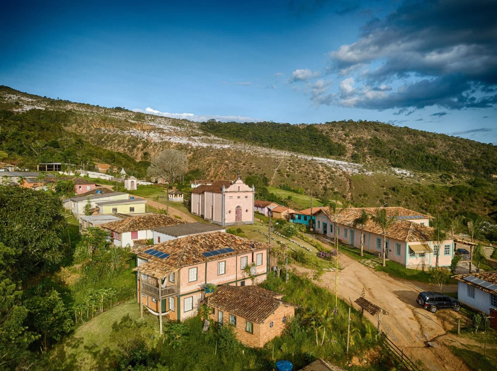 A village that time slowed down in, not stopped. Aerial view of a small historic village in Minas Gerais, with colonial-era pastel-painted houses, terracotta-tiled roofs, a pink chapel, a dirt road, and forested hills with exposed white rock in the background, within the Ibiti Project reserve.