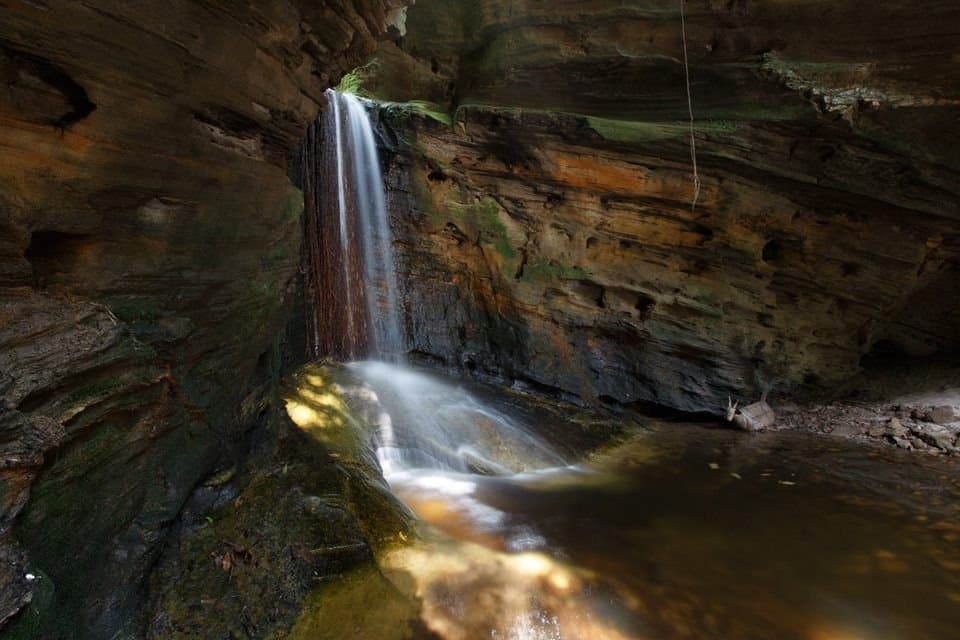 Tucked inside the rock, this waterfall has been keeping its own schedule for a long time. A slender waterfall drops through layered sandstone walls inside a sheltered rocky canyon on the Ibiti Project reserve, pooling in a still, amber-tinted basin below
