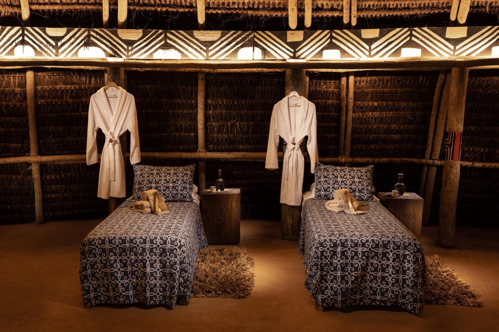 Robes ready, the forest doing the rest of the work. Two massage beds with blue patterned linen and white robes hanging on wooden posts inside a thatched-roof Oca at the Ibiti Project, lit by warm amber light