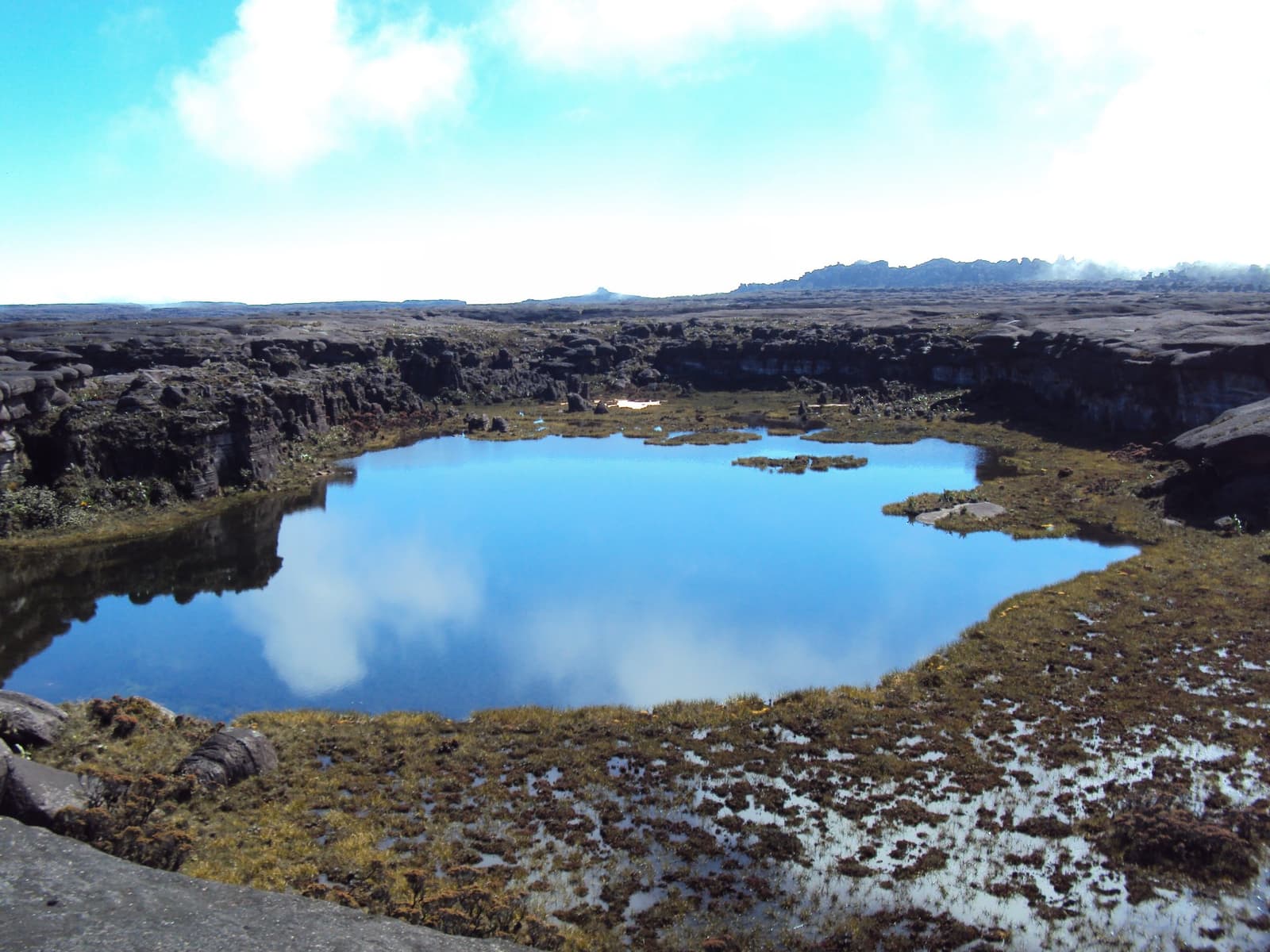 Up here, the sky falls into the rock and the rock doesn't mind. A calm blue reflective pool on the rocky tepui plateau during a mount roraima expedition, surrounded by dark sandstone outcrops, golden mosses, and low clouds mirrored on the water's surface