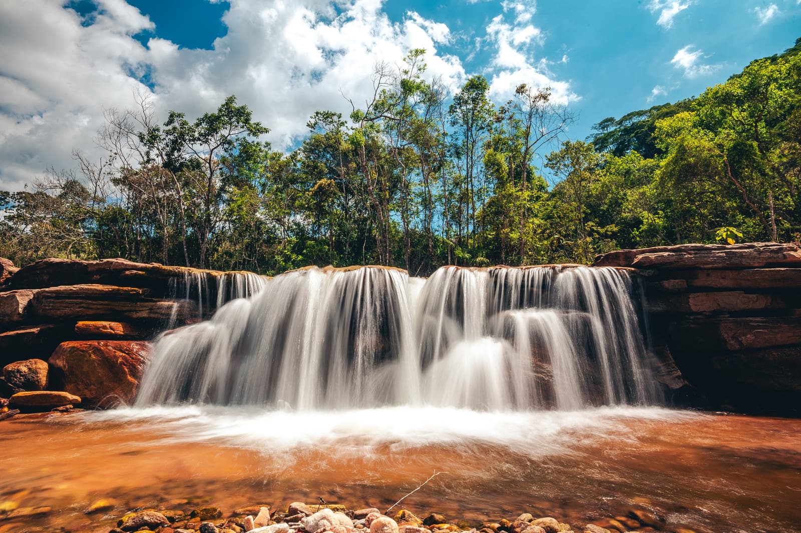Red rocks, white water, and nobody else in sight. Photo: Igor Tibirica A wide waterfall cascading over red sandstone rocks into a copper-toned pool, surrounded by green Atlantic Forest trees under a partly cloudy blue sky, seen on the Ibiti Project's Areião excursion in Minas Gerais.