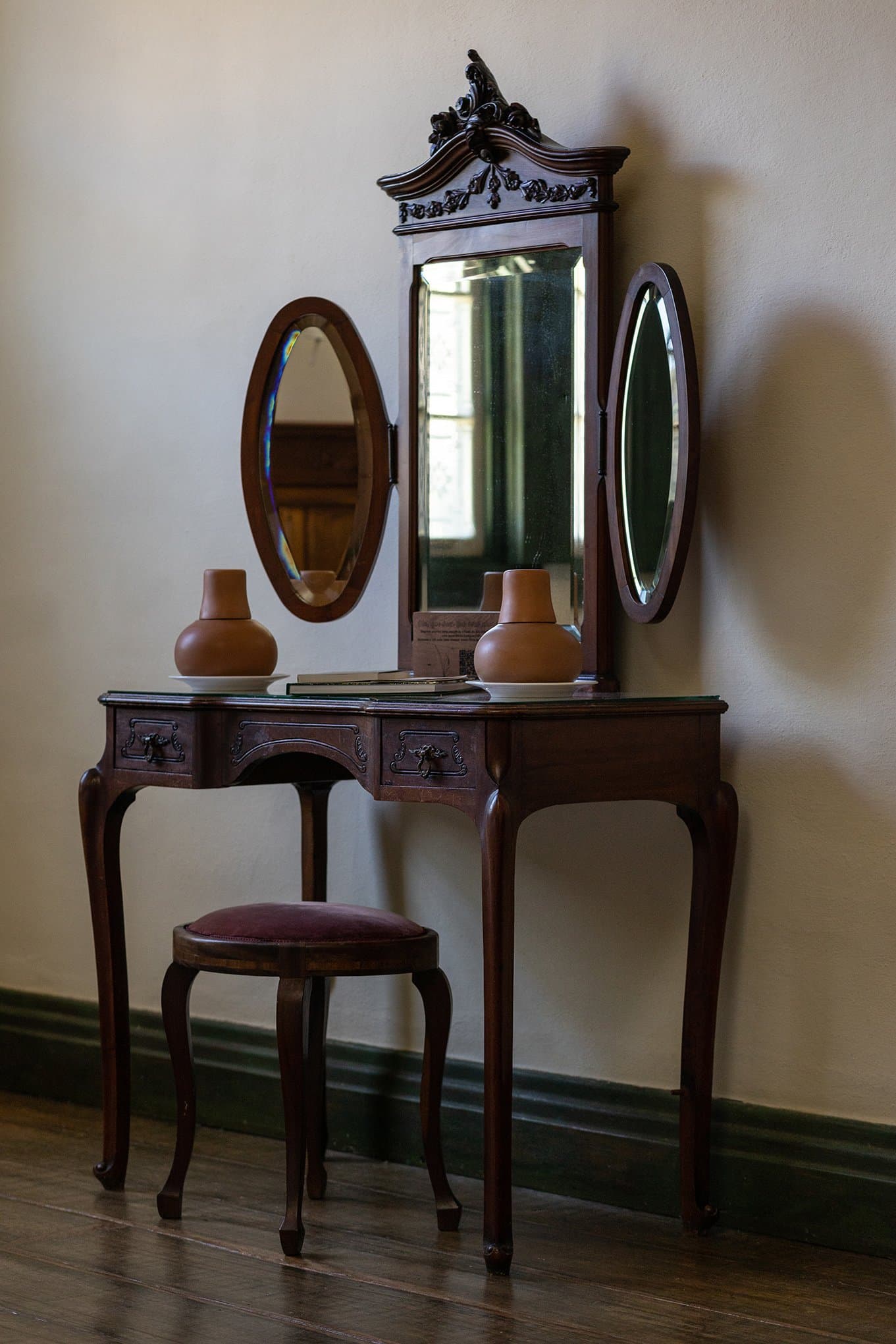 Farmhouse history, still here, holding its own against the forest outside. Photo: Henrique Mangeon Ornate dark-wood vanity table with a carved pediment mirror and two flanking oval mirrors, terracotta vases, and a velvet stool, in an Ibiti Project lodge room