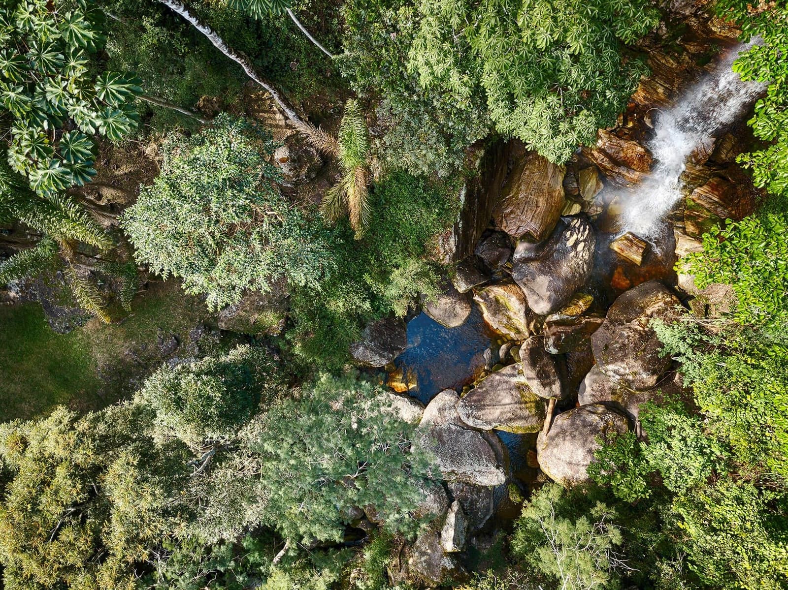 Seen from above, the water finds its own way through. Aerial view of a waterfall cascading over large rounded boulders into a dark natural pool, surrounded by dense Atlantic forest canopy in the Ibiti Project reserve