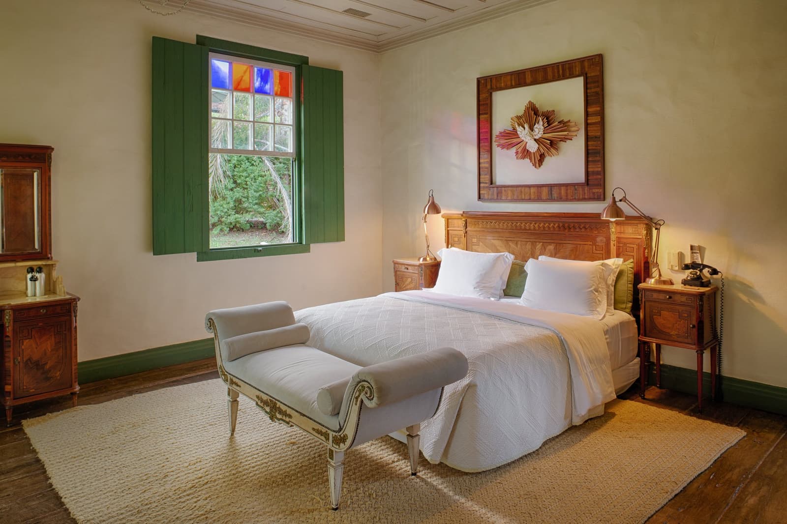 Colonial bones, fresh linen, and a window that keeps the forest close. Photo: Henrique Mangeon A double bedroom at the Ibiti Project's Engenho Lodge, featuring an ornate antique wooden headboard, crisp white linen, copper bedside lamps, a colonial chaise bench, green shuttered window with coloured glass panels, and a framed wall artwork above the bed