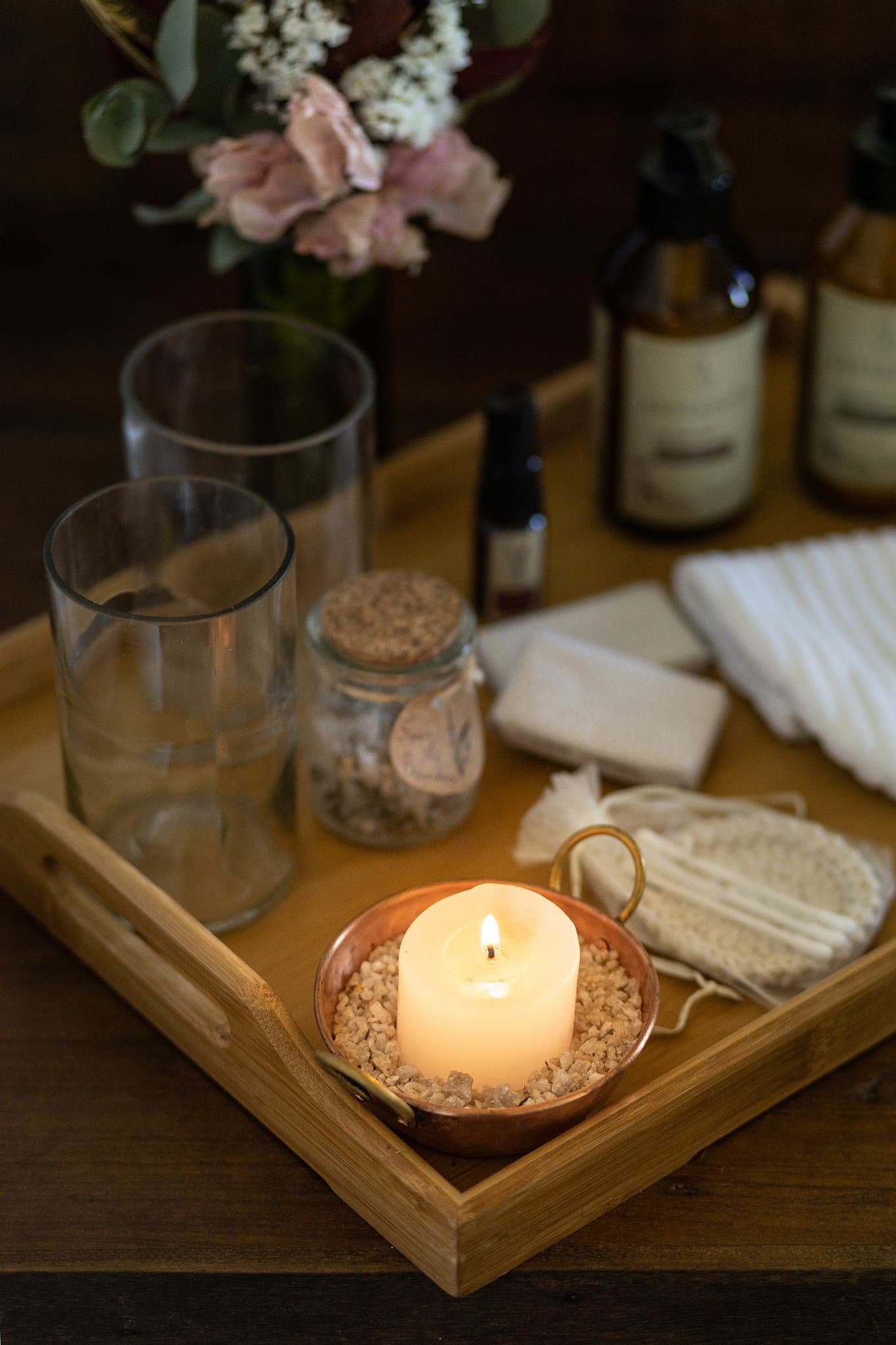 The details that make you forget you had a packed schedule tomorrow. Photo: Henrique Mangeon A lit pillar candle in a copper dish sits on a wooden tray at the Ibiti Project lodge, alongside glass tumblers, a cork-lidded jar of bath salts, a loofah, linen towels, a bar of soap, and amber glass toiletry bottles, with pink flowers softly blurred in the background.