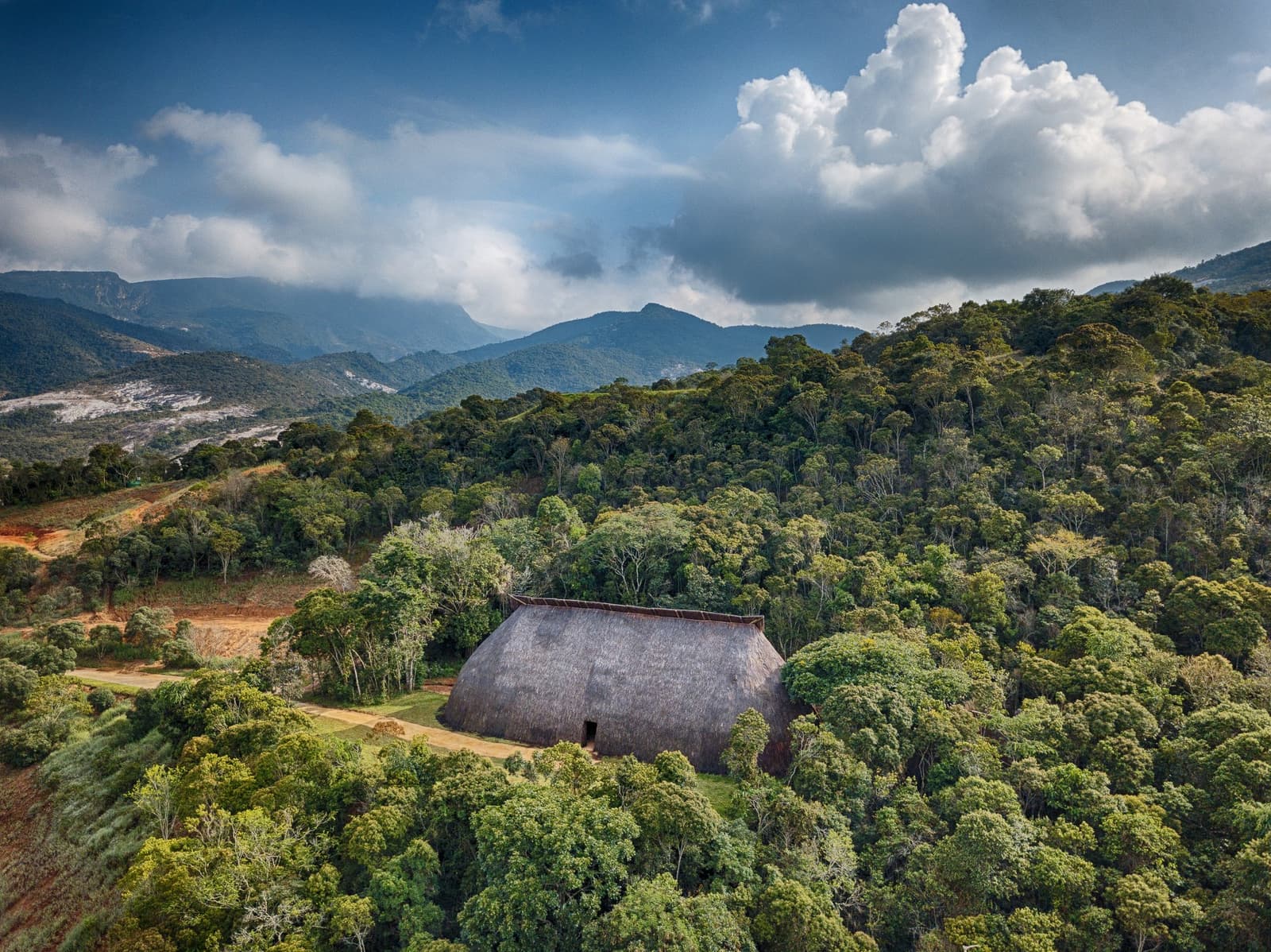 Six thousand hectares of forest, and one thatched roof holding its own. Aerial view of a large traditional thatched-roof Oca structure nestled among dense Atlantic forest trees, with forested mountains and dramatic cumulus clouds in the background at the Ibiti Project in Minas Gerais