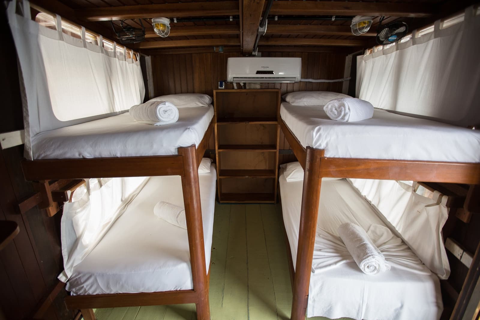 Four bunks, fresh towels, and the Rio Negro right outside the curtain. Photo: sitah Interior of an air-conditioned amazon river cruise brazil cabin with two sets of wooden bunk beds, crisp white linens, rolled towels, curtained portholes, and a central storage unit beneath a timber ceiling