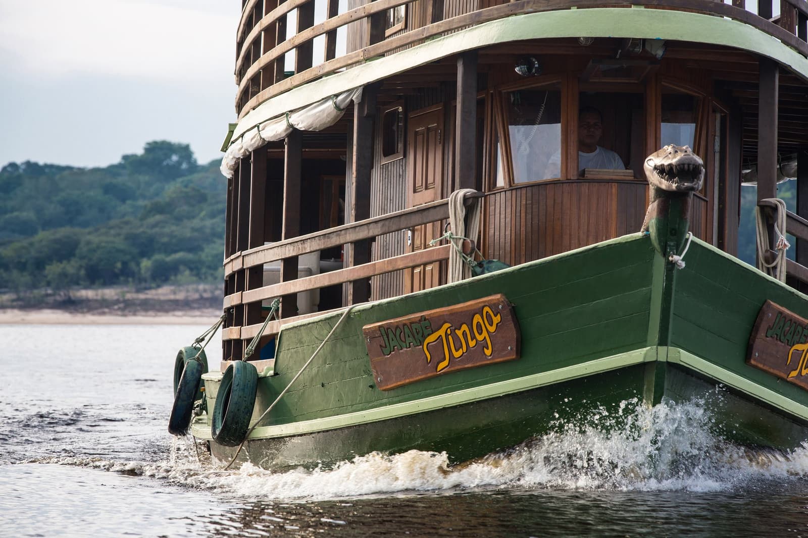 The Jacaré-Tinga knows these rivers, and it shows. Photo: sitah The green wooden bow of the Jacaré-Tinga boat cutting through dark river water on an amazon jungle cruise brazil, with its multi-deck timber superstructure and a carved caiman figurehead visible at the prow