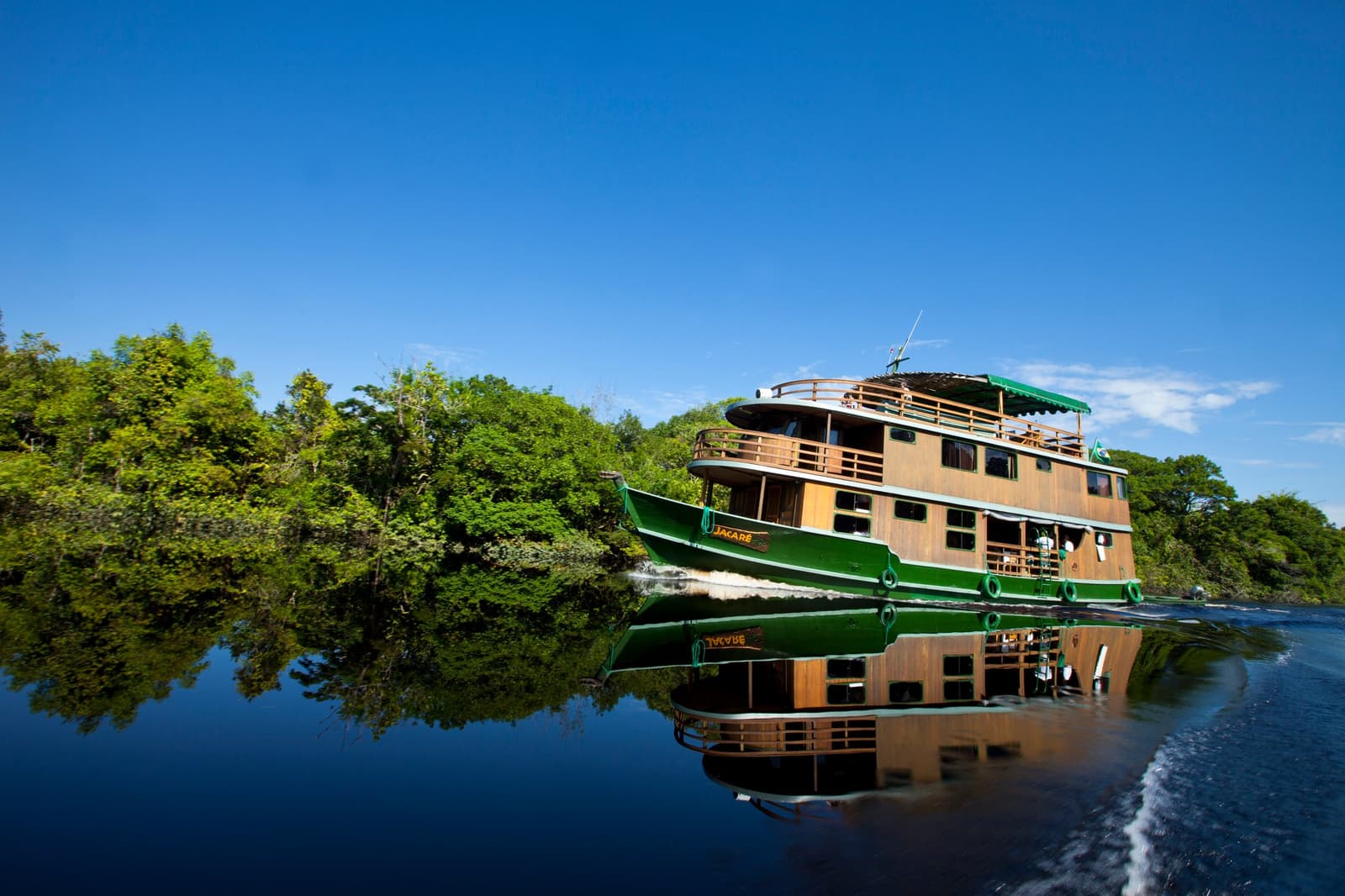 Home for five days, and it moves. Not bad at all. The wooden riverboat Jacaré cruising a dark calm river on an amazon river cruise in Brazil, its green hull perfectly mirrored in the still water with dense rainforest lining both banks under a deep blue sky